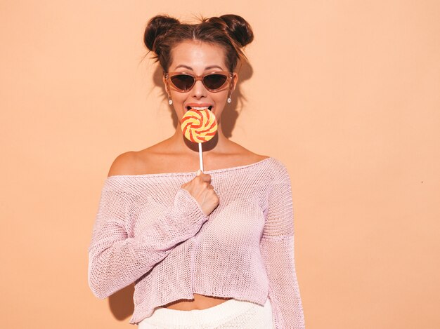 Close-upportret van jonge mooie sexy glimlachende vrouw met lijkenetende kapsel. Trendy meisje in casual zomer kleding in zonnebril. Heet model geïsoleerd op beige. Eten, bijten snoep lolly