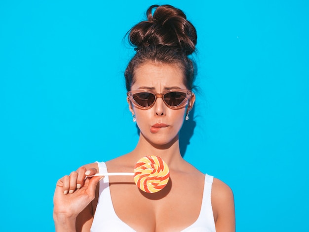 Close-upportret van jonge mooie sexy vrouw met lijkenetende kapsel. Trendy meisje in casual zomer wit zwempak in zonnebril. Heet model geïsoleerd op blauw. Eten, bijten snoep lolly