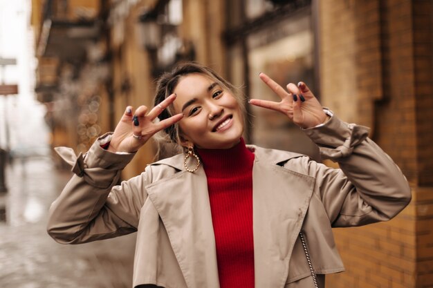 Close-upportret van leuke Aziatische vrouw in rode bovenkant en beige trenchcoat glimlachend en vredestekens op muur van mooi gebouw tonen
