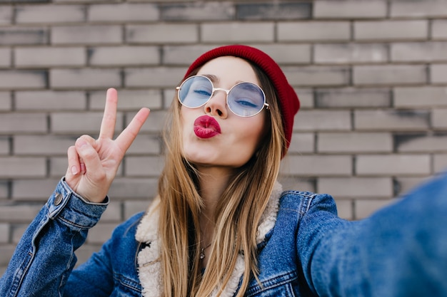 Close-upportret van schitterend wit meisje in ronde glazen die positieve emoties uitdrukken. Fascinerende vrouw selfie maken met kussen gezicht expressie op bakstenen muur.