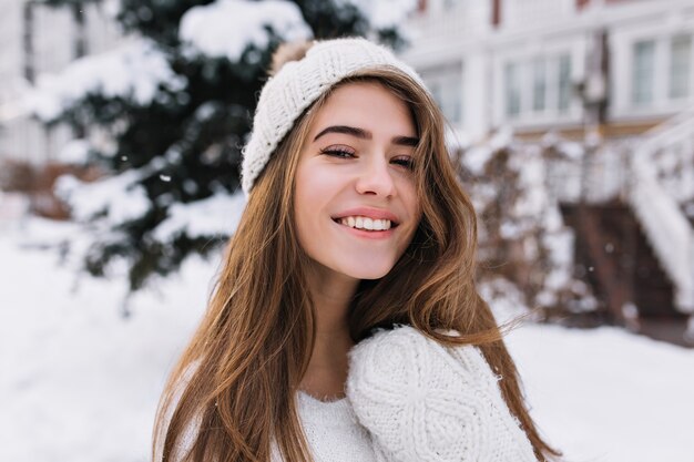 Close-upportret van tevreden blonde vrouw met oprechte glimlach die van winterochtend geniet. Mooie europese vrouw in witte hoed kijken naar besneeuwde weergave buiten.