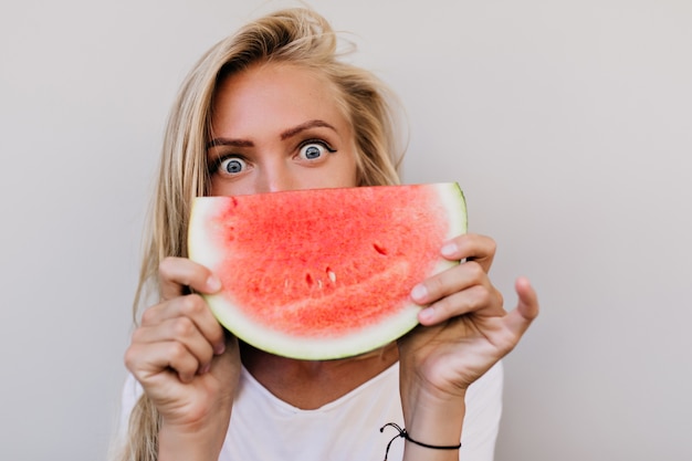 Close-upportret van vrouw met grijze ogen die watermeloen eten. Binnenfoto van spectaculaire blanke dame met lang kapsel die van fruit geniet.