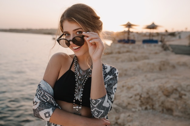 Closeup portret van aantrekkelijk jong meisje stijlvolle zonnebril opstijgen, op zonsondergang, op strand met sensuele look. Trendy zwart topje, ketting, vest met ornamenten. Resort, vakantie, vakantie.