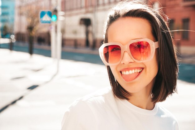 Closeup portret van jonge mooie lachende hipster vrouw in trendy zomerkleren. Sexy zorgeloze vrouw die zich voordeed op de straat achtergrond bij zonsondergang. Positief model toont tong .Gelukkig en vrolijk