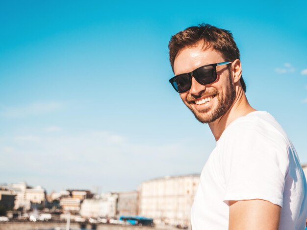 Closeup portret van knappe lachende hipster lamberseksueel modelstijlvolle man gekleed in het wit Tshirt Fashion man poseren achter blauwe hemel op de straat achtergrond in zonnebril