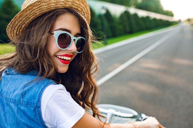 Closeup portret van schattig meisje met lang krullend haar in hoed fietsen op de weg. Ze draagt een jerkin, blauwe zonnebril. Ze lacht naar de camera, uitzicht vanaf de achterkant.