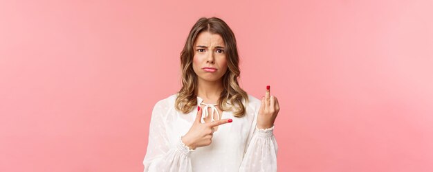 Closeup portret verdrietig en gefrustreerd jong blond meisje wachtte op voorstel tijdens romantische date wijzend op de vinger zonder weddint ring met verbaasde verstoorde uitdrukking staande roze achtergrond