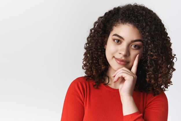 Closeup stijlvolle zelfverzekerde knappe tedere jonge mixedrace vrouw donker krullend kapsel mager hoofd wijsvinger grijnzende opgetogen nieuwsgierige blik doordachte camera interesse gesprek