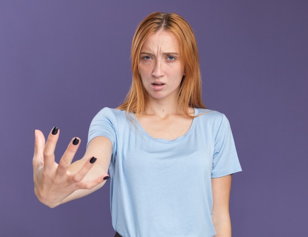 Clueless jong roodharig gembermeisje met sproeten houdt hand open geïsoleerd op paarse muur met kopieerruimte