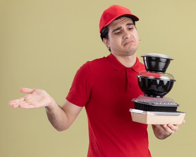 Clueless jonge blanke bezorger in rood uniform en pet die voedselcontainers en papieren voedselpakket vasthoudt en kijkt, ik weet het niet gebaar geïsoleerd op olijfgroene muur