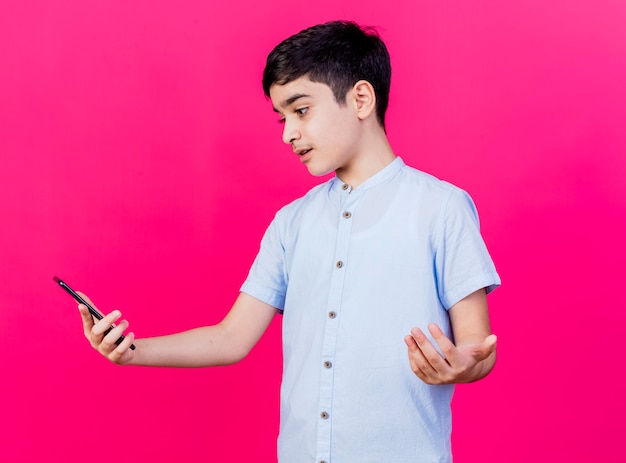 Clueless jonge blanke jongen houden en kijken naar mobiele telefoon met lege hand geïsoleerd op karmozijnrode achtergrond