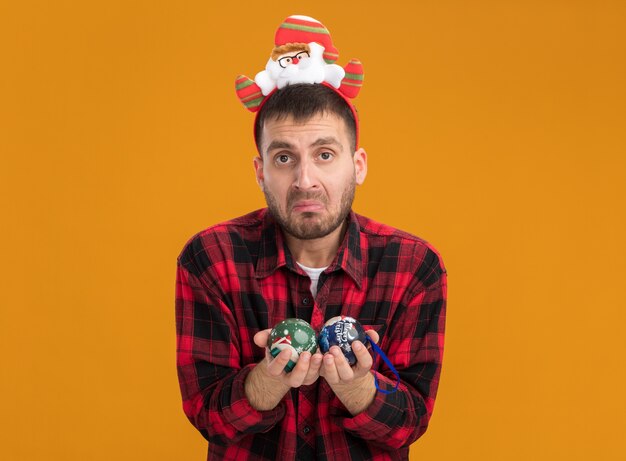 Clueless jonge blanke man met de hoofdband van de kerstman met kerstballen geïsoleerd op oranje muur met kopie ruimte