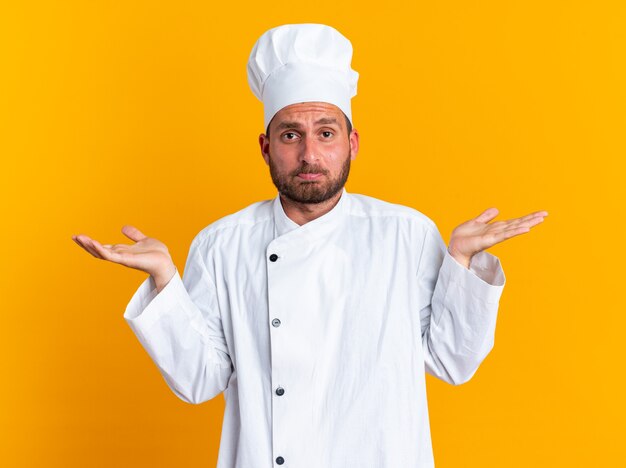 Clueless jonge blanke mannelijke kok in chef-kok uniform en pet kijken camera doen ik weet niet gebaar geïsoleerd op oranje muur
