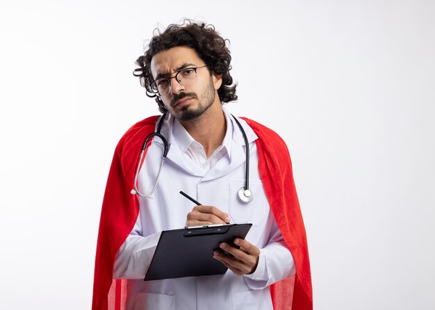 Clueless jonge blanke superheld man in optische bril dragen dokter uniform met rode mantel en met stethoscoop om nek houdt potlood en klembord