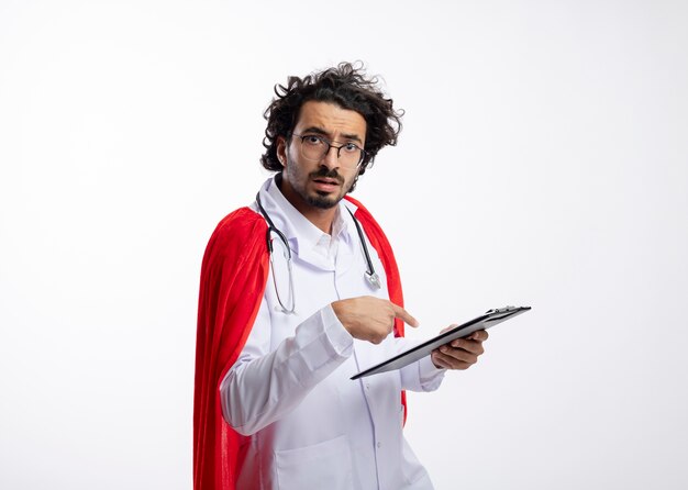 Clueless jonge blanke superheld man in optische bril dragen dokter uniform met rode mantel en met stethoscoop rond nek houdt vast en wijst naar klembord
