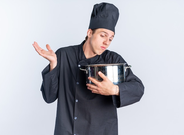 Clueless jonge blonde mannelijke kok in chef-kok uniform en pet houden en kijken in pot met lege hand