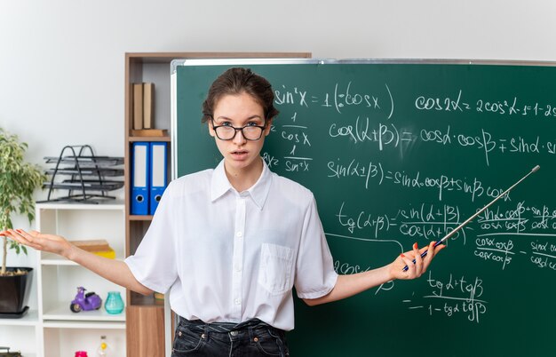 Clueless jonge blonde vrouwelijke wiskundeleraar met een bril die voor een schoolbord staat met een aanwijzer met een lege hand in de klas