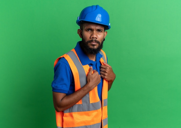 Gratis foto clueless jonge bouwer man in uniform met veiligheidshelm kijkend naar voorkant geïsoleerd op groene muur met kopieerruimte