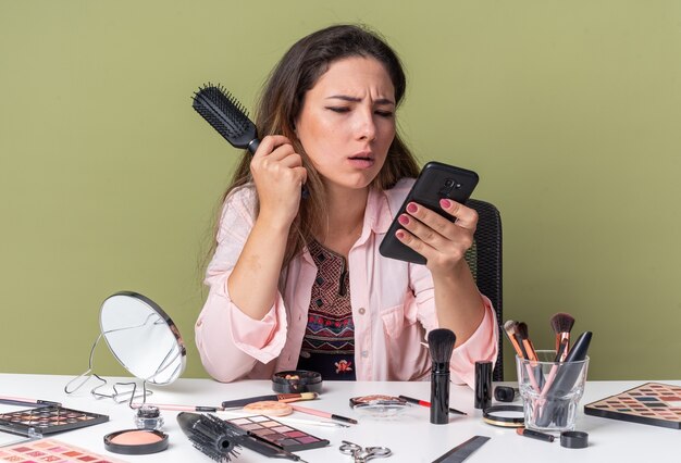 Clueless jonge brunette meisje zittend aan tafel met make-up tools houden kam en kijken naar telefoon geïsoleerd op olijf groene muur met kopie ruimte