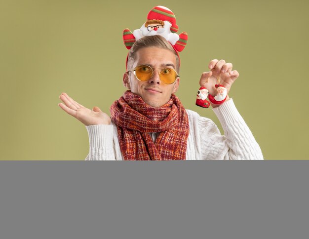 Clueless jonge knappe kerel die de hoofdband en de sjaal van de Kerstman draagt ?? die kerstversieringen van de Kerstman houdt die lege hand tonen die op olijfgroene muur wordt geïsoleerd