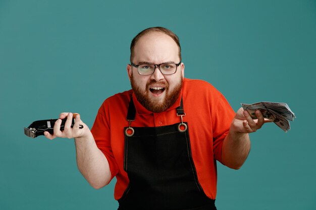 Clueless jonge mannelijke kapper dragen van een bril rood shirt en kapper schort kijken camera met geld en tondeuse in handen geïsoleerd op blauwe achtergrond