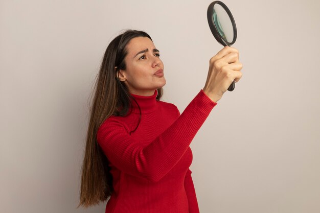 Clueless mooie vrouw houdt en kijkt naar vergrootglas geïsoleerd op een witte muur