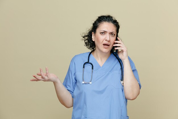 Clueless vrouwelijke arts van middelbare leeftijd, gekleed in uniform en stethoscoop om de nek, kijkend naar camera die lege hand toont terwijl hij aan de telefoon praat geïsoleerd op olijfgroene achtergrond