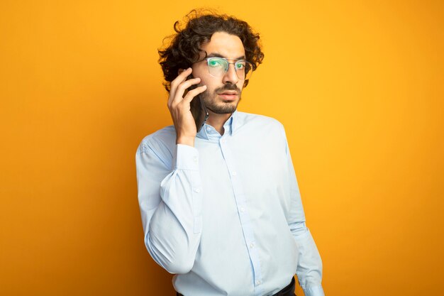 Cofident jonge knappe blanke man met bril praten over de telefoon kijken camera geïsoleerd op een oranje achtergrond met kopie ruimte