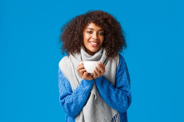 Comfort, tederheid en warmte concept. Charmante mooie Afro-Amerikaanse vrouw met afro kapsel, in trui en sjaal, opwarmen met lekker kopje thee, genieten van warme koffie.