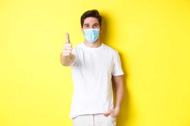 Concept van coronavirus, pandemie en sociale afstand nemen. Zelfverzekerde jonge man in medisch masker duimen opdagen, gele achtergrond.
