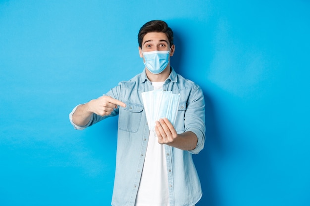 Concept van coronavirus, quarantaine en sociale afstand. Jonge man wijzend op medische maskers, maatregelen tegen covid-19 voorkomend, staande over blauwe achtergrond