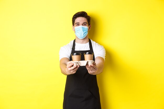 Concept van covid-19, café en sociaal afstand nemen. Barista in medisch masker serveert koffie in meeneembekers, staande in zwarte schort tegen gele achtergrond.
