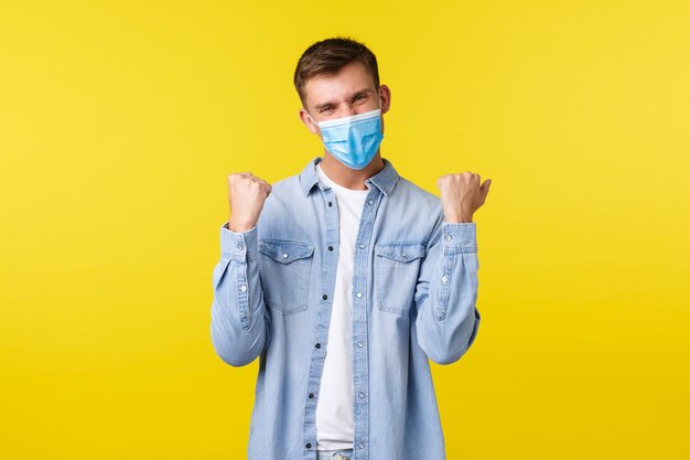 Concept van covid-19 pandemische uitbraak, levensstijl tijdens sociale afstand van het coronavirus. Gelukkige knappe man met medisch masker, vuistpomp en ja zeggen, zich verheugen over overwinning, geluk voelen.