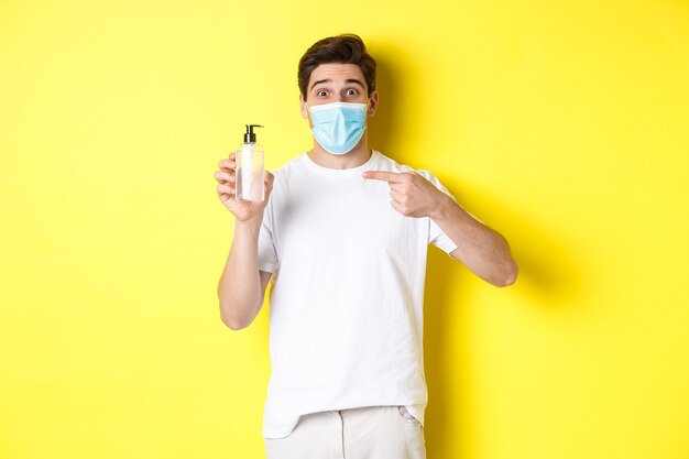 Concept van covid-19, quarantaine en levensstijl. Opgewonden man in medisch masker met goed handdesinfecterend middel, wijzende vinger op antisepticum, staande op gele achtergrond.