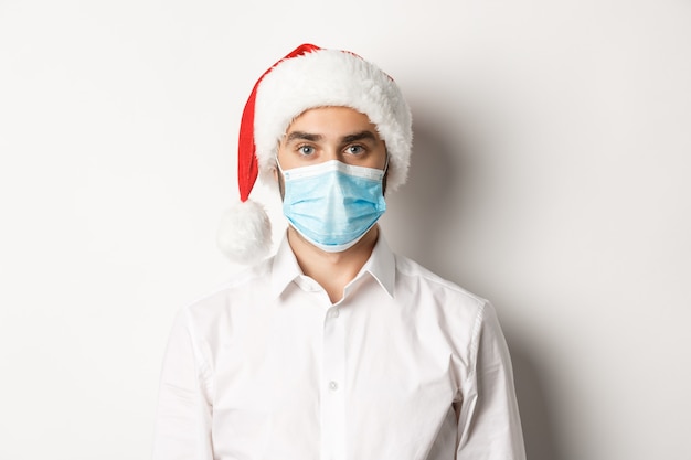 Concept van covid-19, sociaal afstand nemen en wintervakantie. Close-up van jonge man met kerstmuts en gezichtsmasker van coronavirus, Nieuwjaar vieren