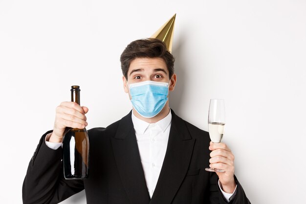 Concept van feest tijdens covid-19. Close-up van knappe man in pak, grappige hoed en medisch masker, fles champagne vasthoudend, nieuwjaar vierend tijdens coronavirus