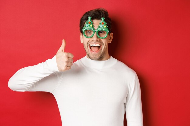Concept van Kerstmis, wintervakantie en feest. Opgewonden knappe man in feestbril, geamuseerd glimlachend en duim omhoog, staande over rode achtergrond.