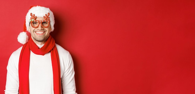 Concept van Kerstmis, wintervakantie en feest. Vrolijke knappe jongen in nieuwjaarsfeestbril en glimlachend, staande over rode achtergrond.