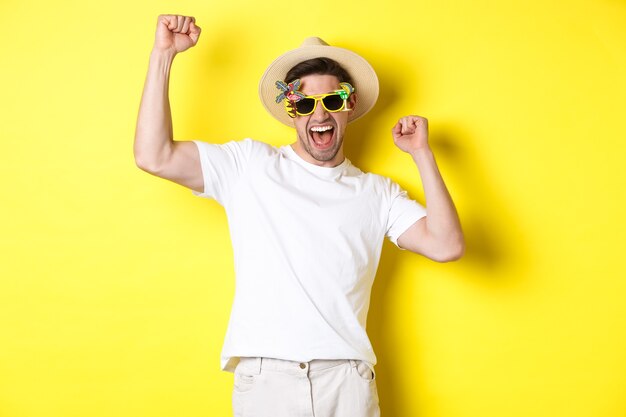 Concept van toerisme en levensstijl. Gelukkige gelukkige kerel die reis wint, zich verheugt en vakantie-outfit, zomerhoed en zonnebril draagt, gele achtergrond.