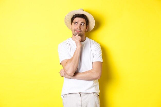 Concept van toerisme en zomer. Doordachte man toerist nadenken, kijken naar de linker bovenhoek en denken, staande in stro hoed en wit t-shirt tegen gele achtergrond.