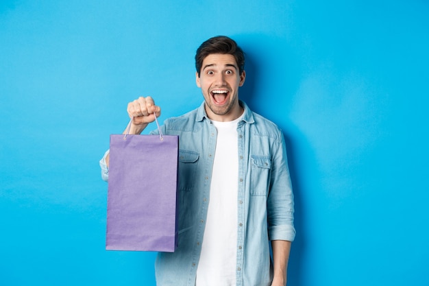 Concept van winkelen, vakantie en levensstijl. Opgewonden knappe kerel die een papieren zak met cadeau vasthoudt en er gelukkig uitziet, staande over een blauwe achtergrond