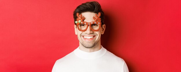 Concept van wintervakantie, kerstmis en feest. Close-up van aantrekkelijke brunette man in feestbril, glimlachend en gelukkig kijkend, staande tegen een rode achtergrond.