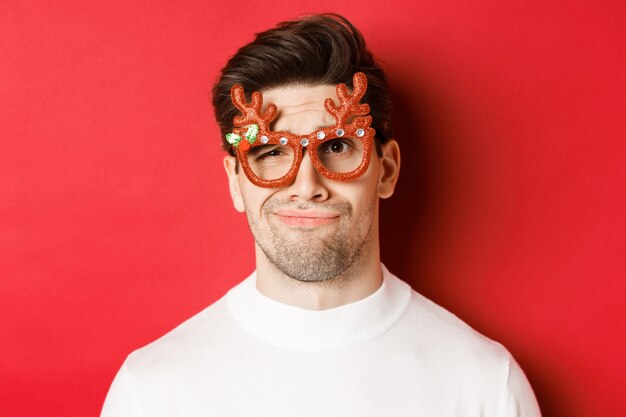 Concept van wintervakantie, kerstmis en feest. Close-up van een sceptische knappe man met een feestbril, die er twijfelachtig en ongeamuseerd uitziet, staande tegen een rode achtergrond