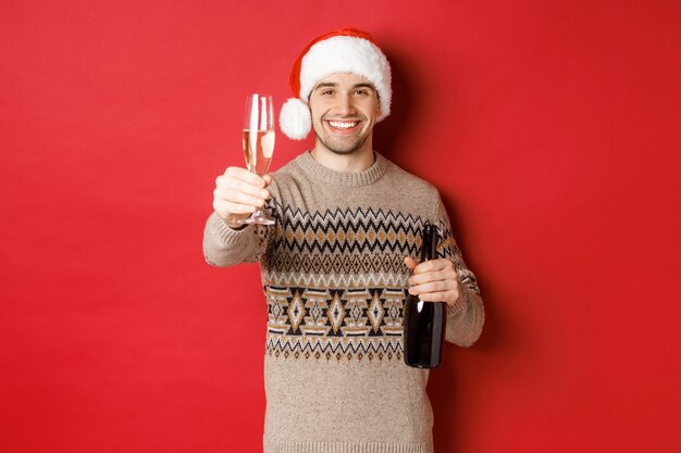 Concept van wintervakantie, nieuwjaar en feest. Portret van een knappe man in kerstmuts en trui, champagne vasthoudend, glas heffend en gejuich zeggen op kerstfeest