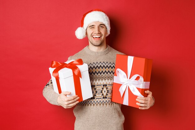 Concept van wintervakantie, nieuwjaar en feest. Portret van een zelfverzekerde en brutale jongeman bereidde cadeaus voor Kerstmis, knipoogde en hield cadeautjes vast, staande op een rode achtergrond