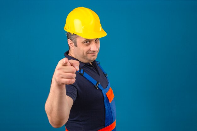 Constructor man van middelbare leeftijd met eenvormige bouw en veiligheidshelm die gelukkig glimlacht en zijn wijsvingers op camera over geïsoleerde blauwe muur richt