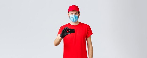 Contactloze leveringsbetaling en online winkelen tijdens covid19 zelfquarantaine Knappe koerier in rode uniforme pet medisch gezichtsmasker en handschoenen tonen creditcardbestelling internet