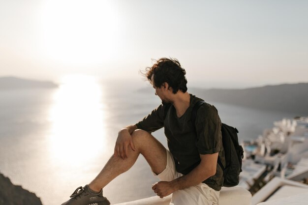 Cool brunette man kijkt naar zee Gebruinde man in witte korte broek en donkergroen t-shirt zit op het hek en geniet van zonsondergang