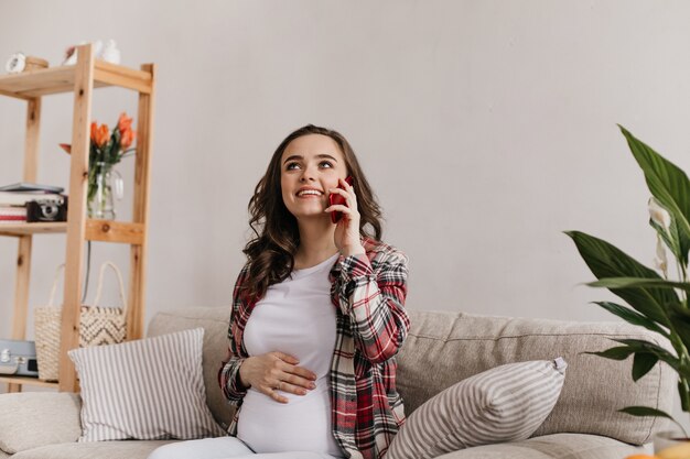 Cool jonge brunette zwangere vrouw in witte tee en geruite overhemd glimlacht en gesprekken op mobiel