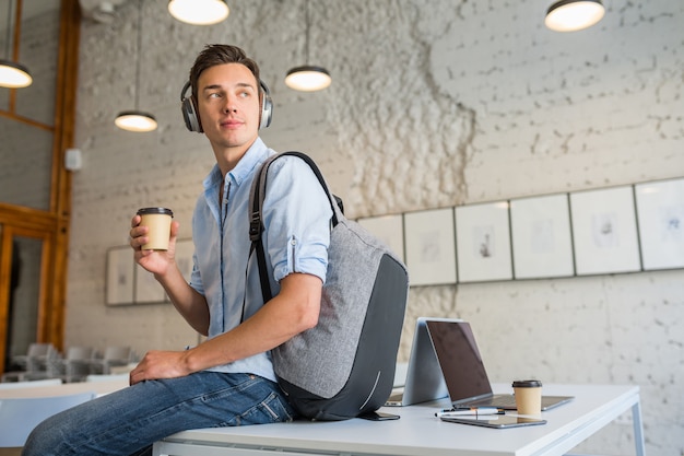 Cool jonge knappe man zittend op tafel in koptelefoon met rugzak in co-working office koffie drinken,